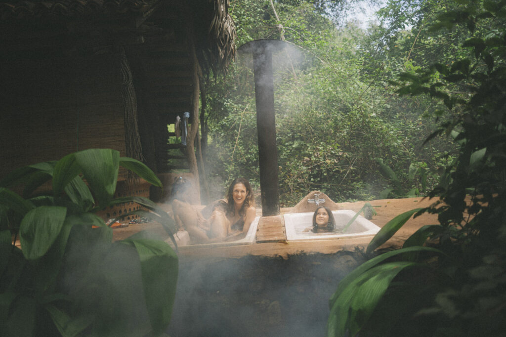 Two children and a parent in a wood-fired outdoor bathtub among broad green leaves, steam rising into the jungle.