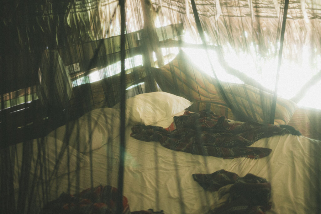A bed under a mosquito net inside a thatched hut, late afternoon sun pouring through the woven roof.