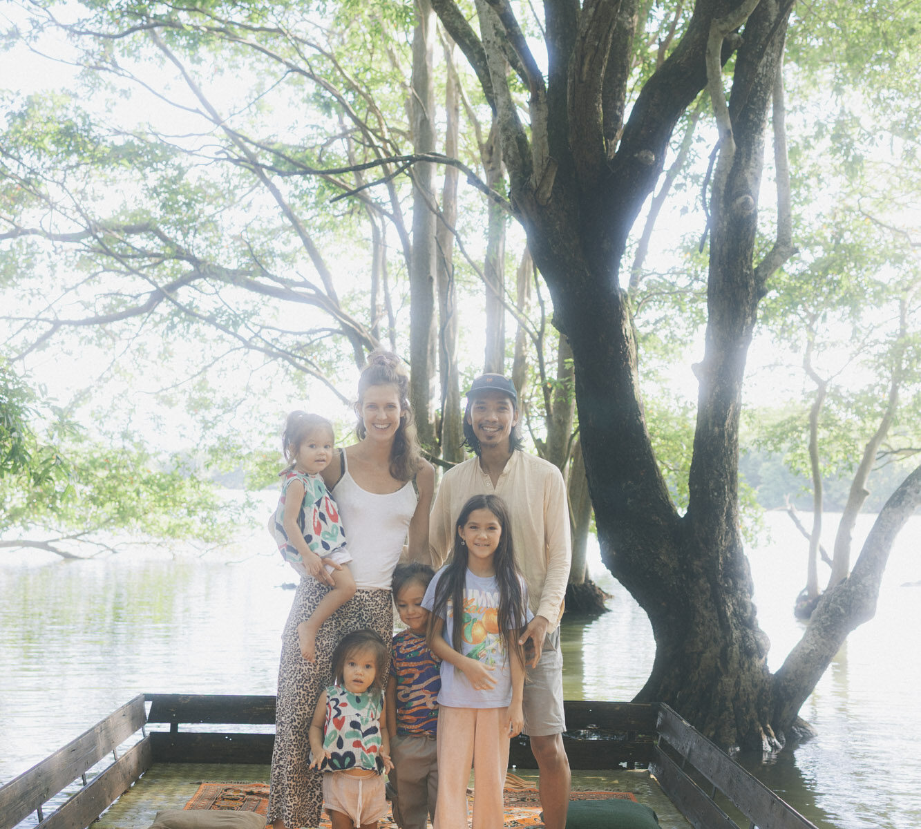 A family of six on a rug at the edge of the lagoon under a wide tree.