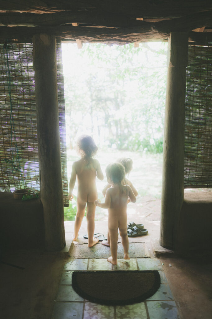 Three small children seen from behind in a low doorway, silhouetted against bright green foliage.