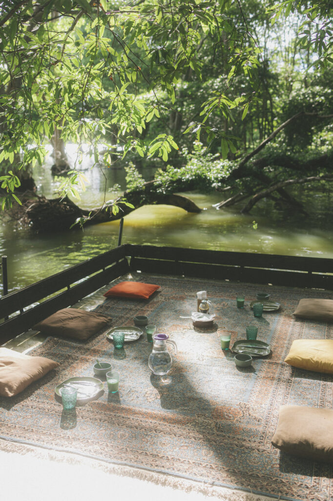 A low picnic set with green glassware on a carpet at the edge of a still lagoon, framed by hanging branches.