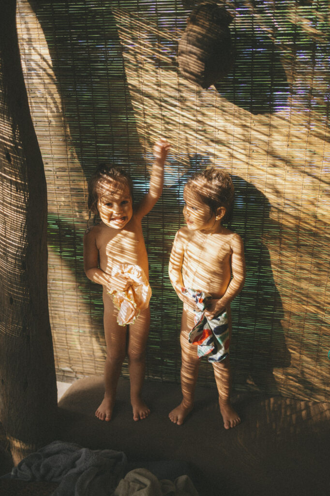 Two toddlers standing against a bamboo blind, sunlight striping their bodies in golden bars.