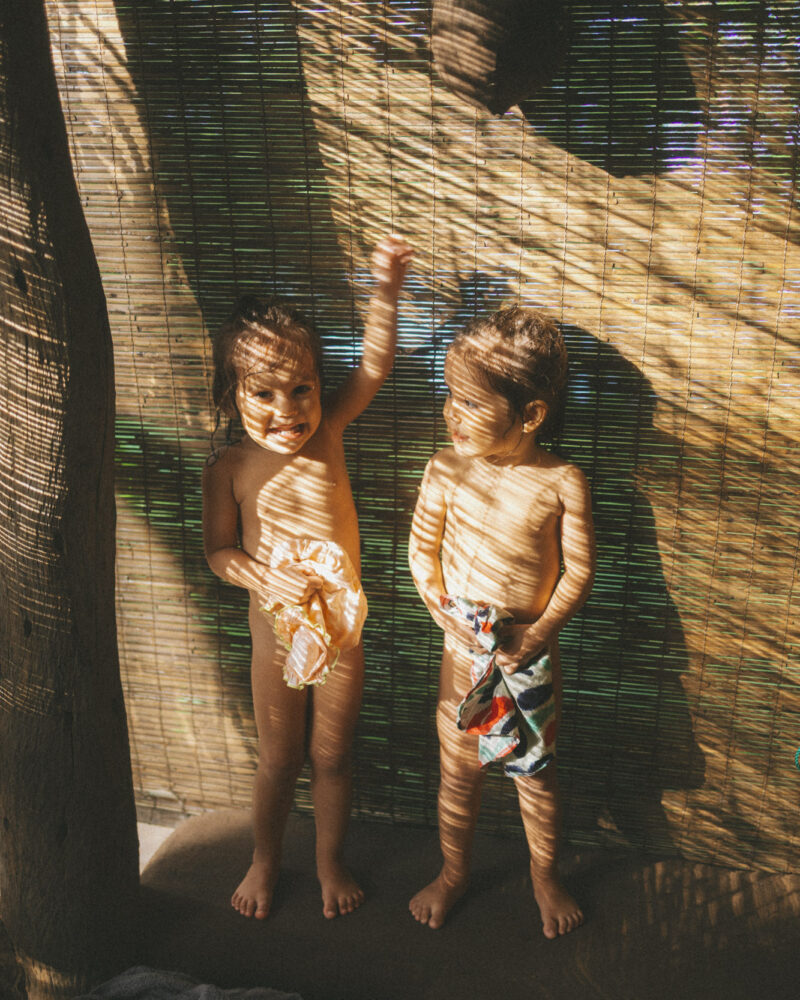Two toddlers standing against a bamboo blind, sunlight striping their bodies in golden bars.